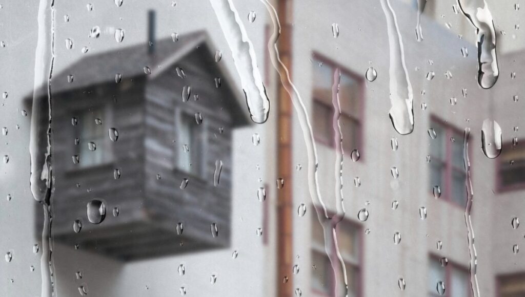 Vista a través de un ventanal donde las gotas de lluvia se deslizan sutilmente, ofreciendo una atmósfera serena y exclusiva. Al fondo, se distingue -aunque suavemente desenfocada- una terraza de madera integrada a una vivienda de tonos claros, reflejo del diseño contemporáneo y elegante propio del sector premium. La escena diurna bajo la llovizna resalta la privacidad y el confort del espacio, atributos ideales para quienes buscan invertir en propiedades excepcionales que combinan sofisticación arquitectónica con ambientes acogedores incluso en días lluviosos.