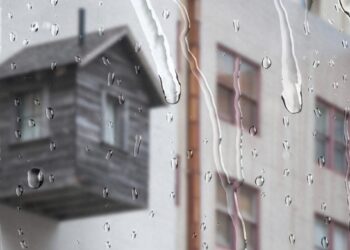 Vista a través de un ventanal donde las gotas de lluvia se deslizan sutilmente, ofreciendo una atmósfera serena y exclusiva. Al fondo, se distingue -aunque suavemente desenfocada- una terraza de madera integrada a una vivienda de tonos claros, reflejo del diseño contemporáneo y elegante propio del sector premium. La escena diurna bajo la llovizna resalta la privacidad y el confort del espacio, atributos ideales para quienes buscan invertir en propiedades excepcionales que combinan sofisticación arquitectónica con ambientes acogedores incluso en días lluviosos.