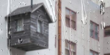 Vista a través de un ventanal donde las gotas de lluvia se deslizan sutilmente, ofreciendo una atmósfera serena y exclusiva. Al fondo, se distingue -aunque suavemente desenfocada- una terraza de madera integrada a una vivienda de tonos claros, reflejo del diseño contemporáneo y elegante propio del sector premium. La escena diurna bajo la llovizna resalta la privacidad y el confort del espacio, atributos ideales para quienes buscan invertir en propiedades excepcionales que combinan sofisticación arquitectónica con ambientes acogedores incluso en días lluviosos.