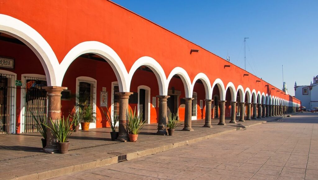 Una elegante hilera de arcos blancos, sostenidos por sólidas columnas en tonos marrones, realza la fachada de esta exclusiva propiedad de color rojo intenso en Puebla, bajo un cielo azul inmaculado. El pasillo se embellece con plantas ornamentales dispuestas estratégicamente y un piso de piedra cuidadosamente colocado, que resalta la autenticidad y el atractivo arquitectónico tradicional. Esta combinación única ofrece una oportunidad inigualable para inversores o compradores que buscan adquirir un inmueble premium con gran valor estético, ubicación privilegiada y potencial para usos residenciales o comerciales exclusivos.