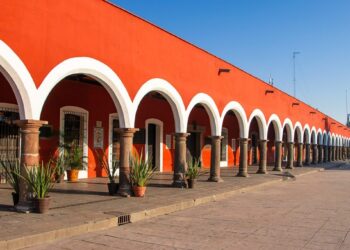 Una elegante hilera de arcos blancos, sostenidos por sólidas columnas en tonos marrones, realza la fachada de esta exclusiva propiedad de color rojo intenso en Puebla, bajo un cielo azul inmaculado. El pasillo se embellece con plantas ornamentales dispuestas estratégicamente y un piso de piedra cuidadosamente colocado, que resalta la autenticidad y el atractivo arquitectónico tradicional. Esta combinación única ofrece una oportunidad inigualable para inversores o compradores que buscan adquirir un inmueble premium con gran valor estético, ubicación privilegiada y potencial para usos residenciales o comerciales exclusivos.