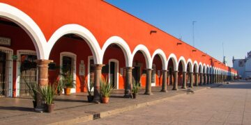Una elegante hilera de arcos blancos, sostenidos por sólidas columnas en tonos marrones, realza la fachada de esta exclusiva propiedad de color rojo intenso en Puebla, bajo un cielo azul inmaculado. El pasillo se embellece con plantas ornamentales dispuestas estratégicamente y un piso de piedra cuidadosamente colocado, que resalta la autenticidad y el atractivo arquitectónico tradicional. Esta combinación única ofrece una oportunidad inigualable para inversores o compradores que buscan adquirir un inmueble premium con gran valor estético, ubicación privilegiada y potencial para usos residenciales o comerciales exclusivos.